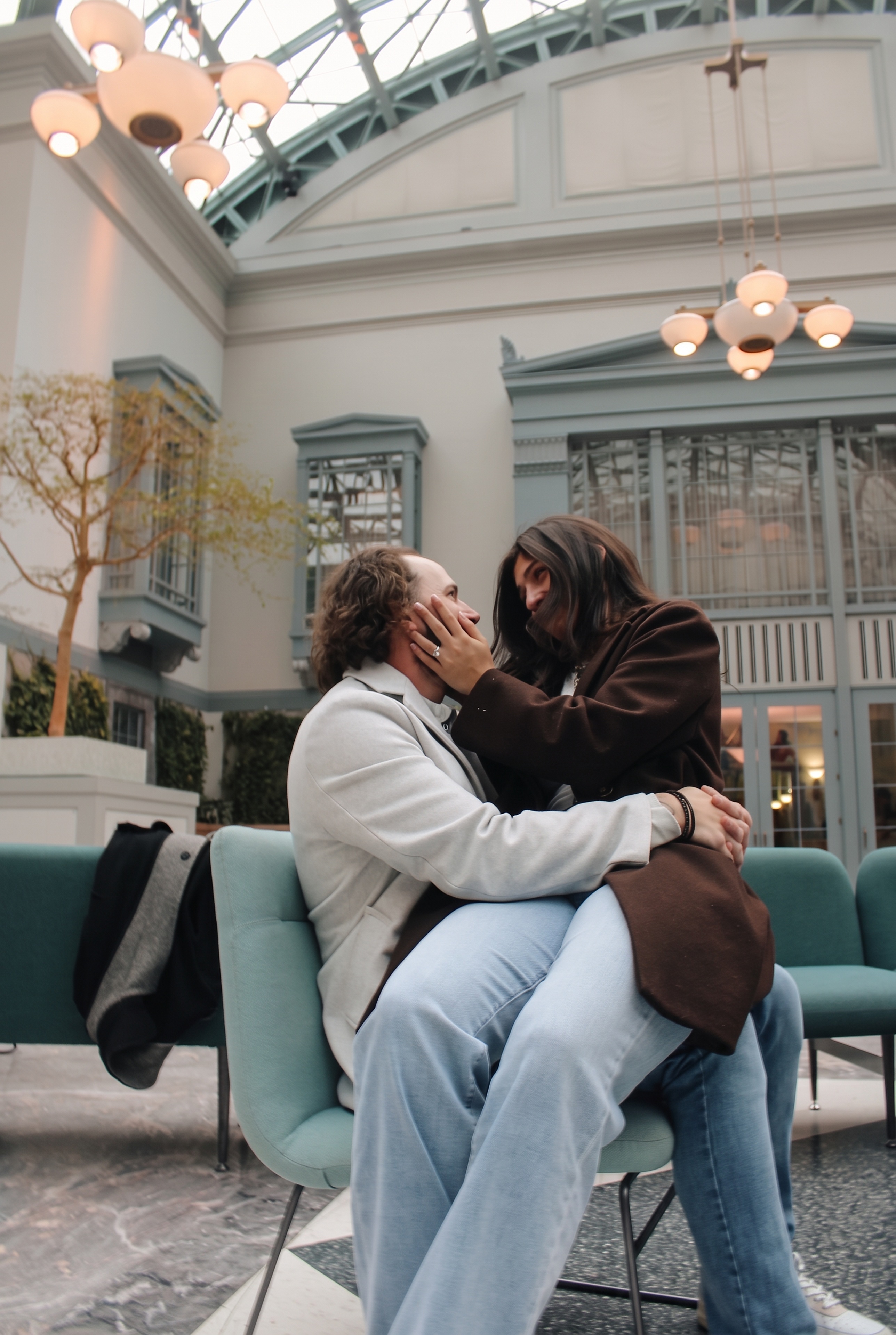Mariella holding Nick's face as they sit together beneath the atrium lights