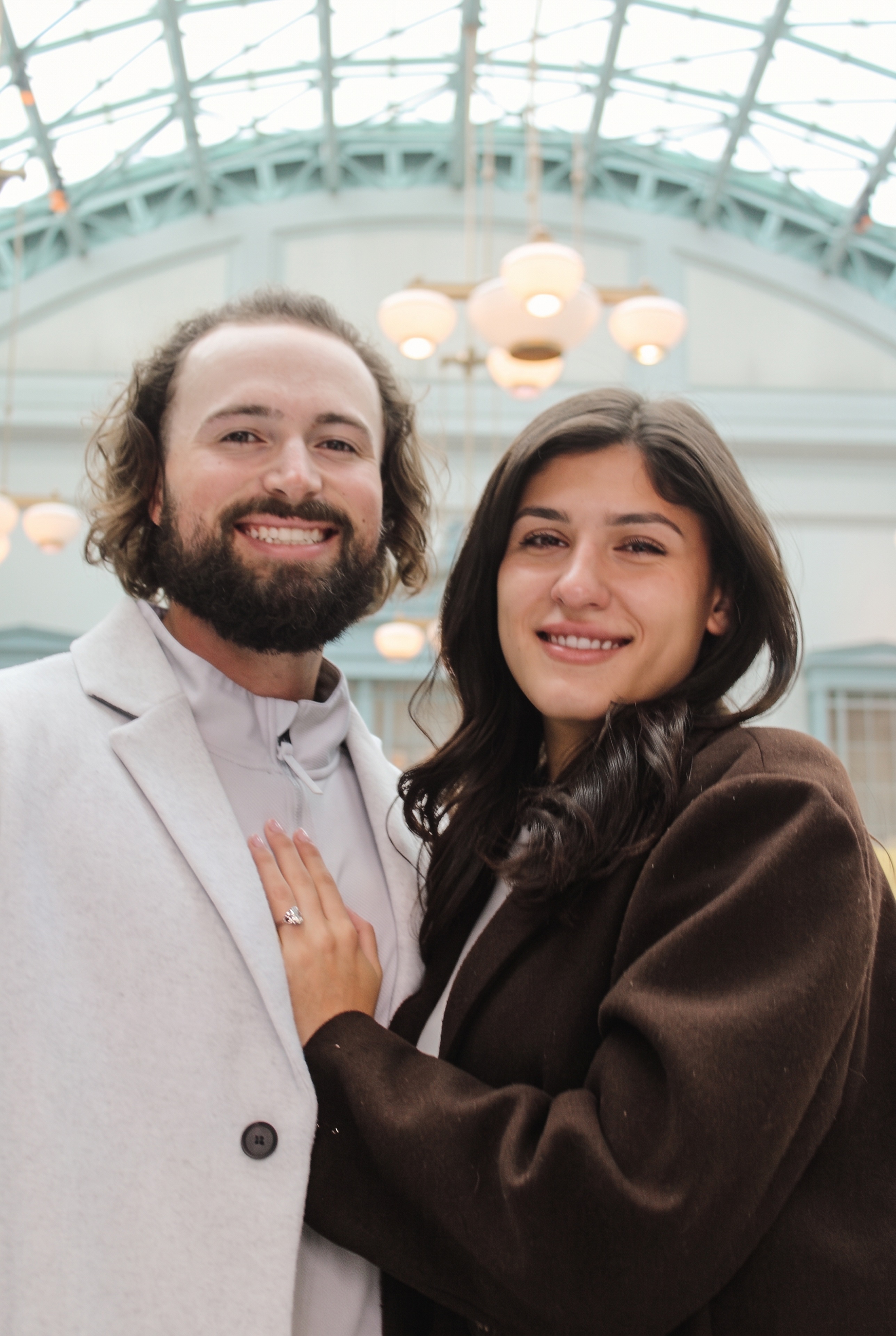 Nick and Mariella smiling beneath the glass atrium, showing her engagement ring