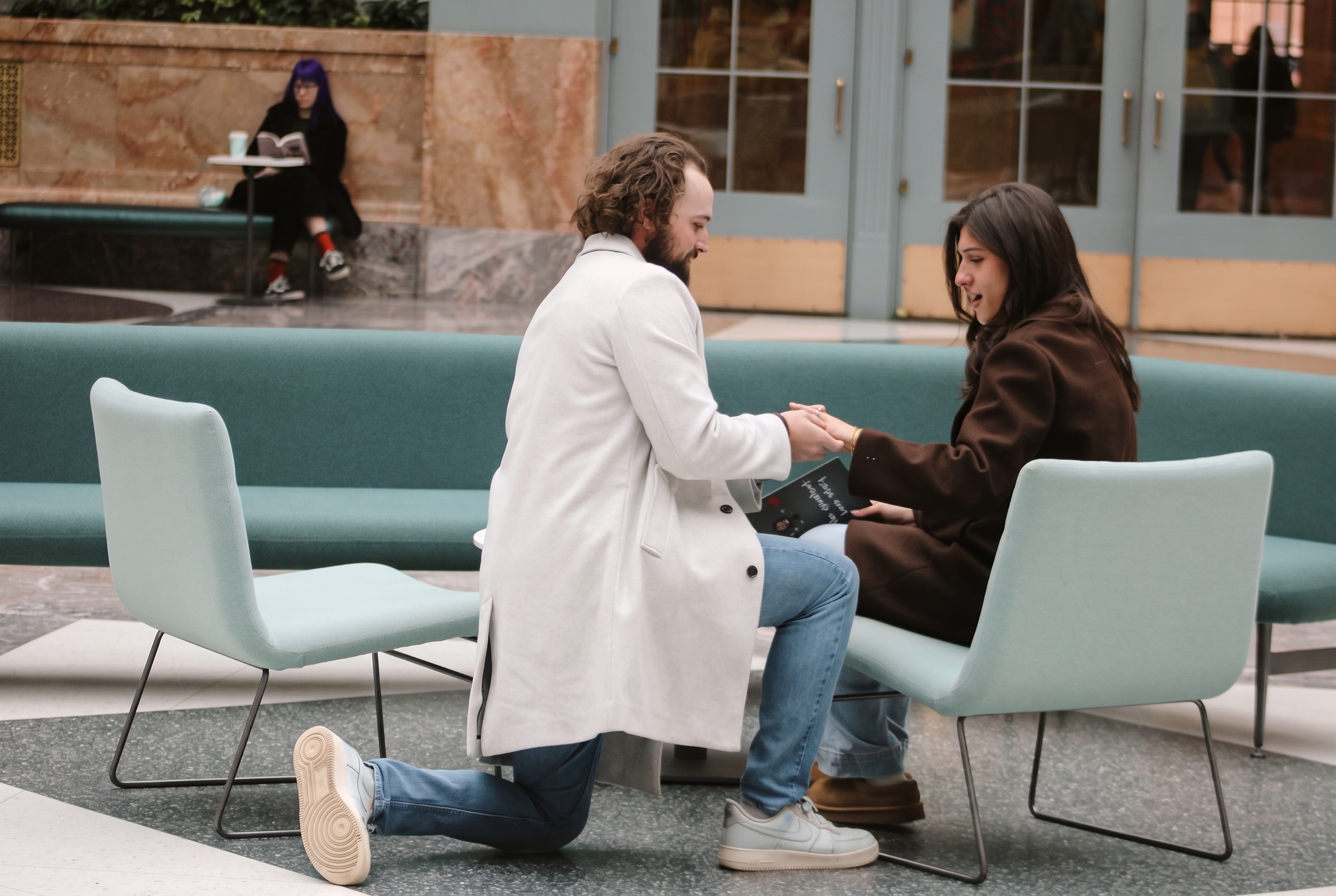 Nick kneeling to propose to Mariella beside the teal lounge chairs