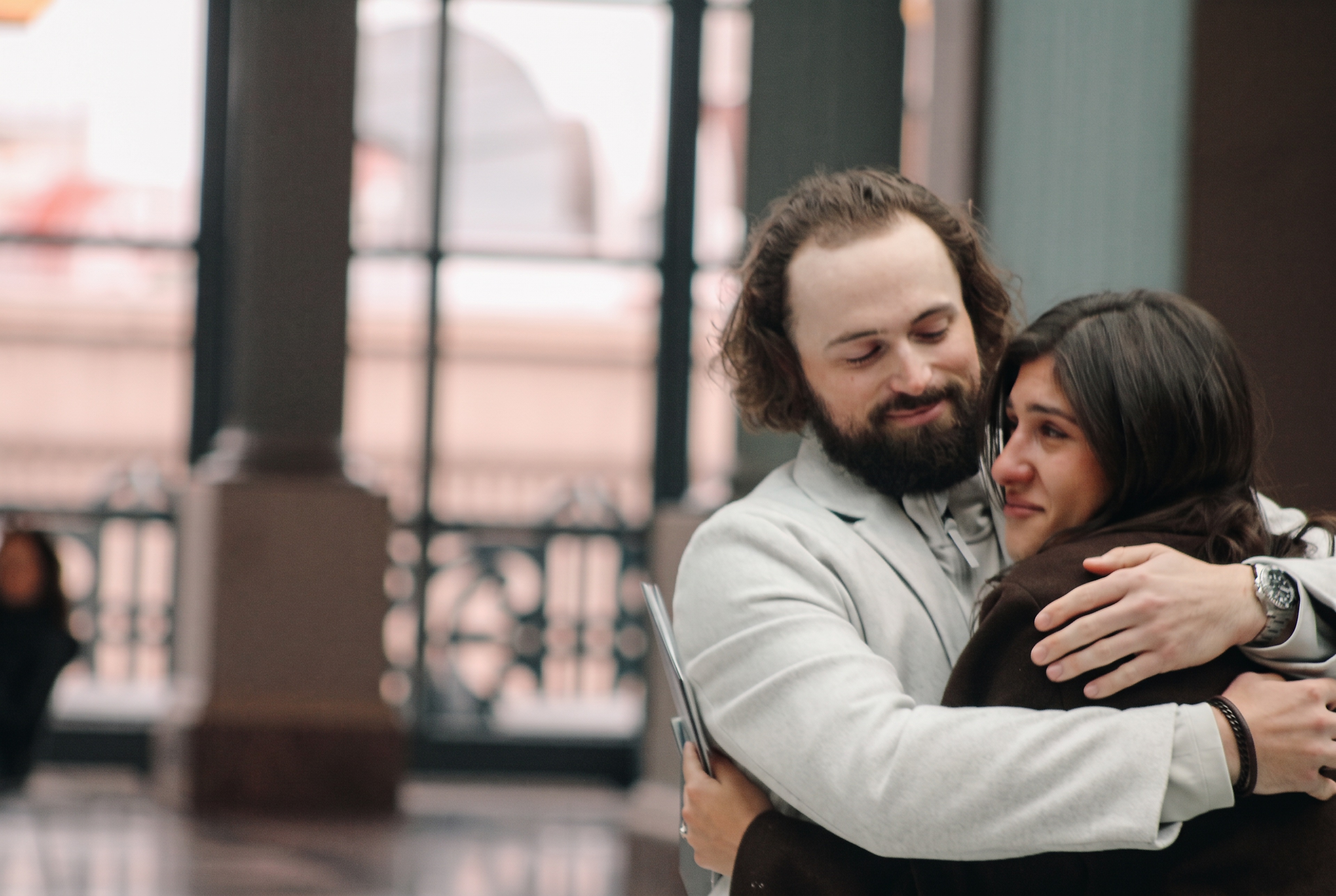 Nick and Mariella embracing by the atrium windows after the proposal
